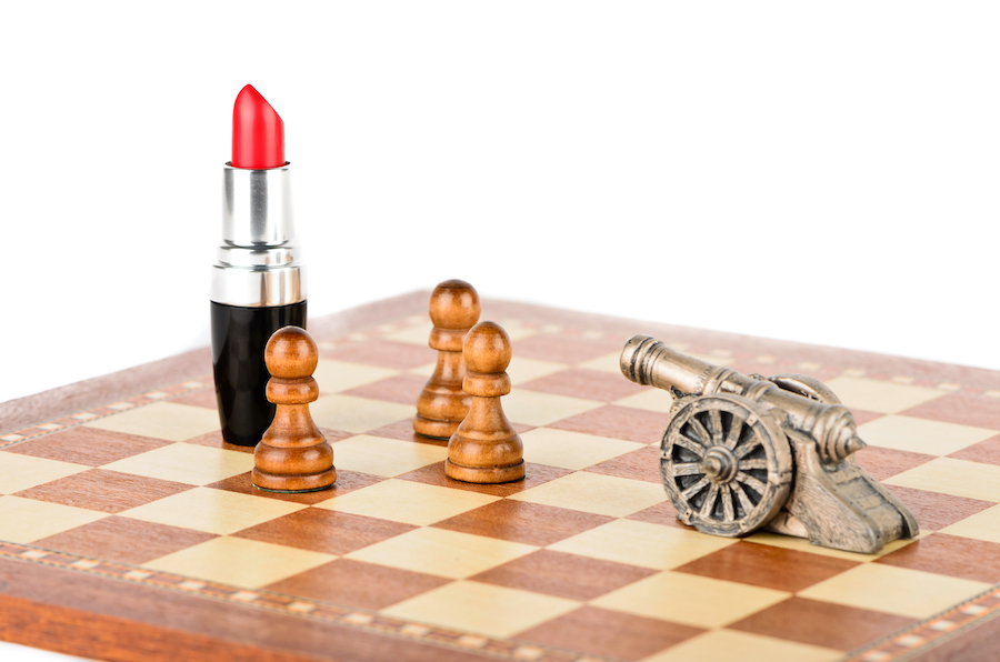 A tube of lipstick sitting on a chess board, being defended from a cannon by three pawns
