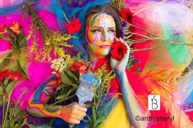 Brightly colored photo of woman holding a paintbrush and flower with vivid face paint