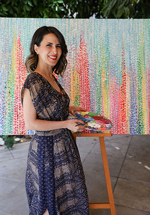 Sheryl Benjy standing in front of a painting on an easel, holding a palette and paint brush