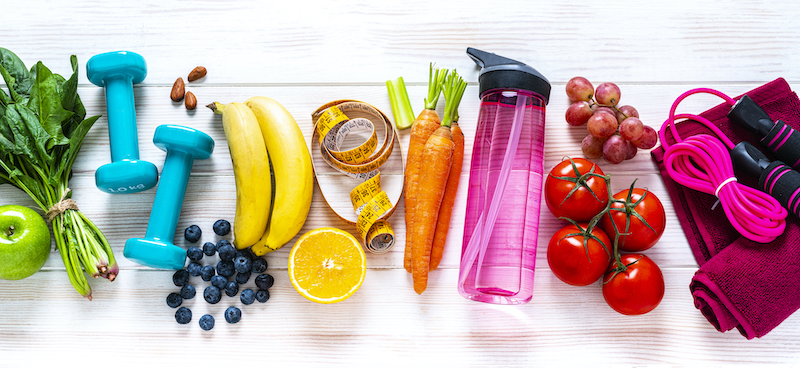 Healthy fruits and vegetables and exercise equipment spread across a white wood table