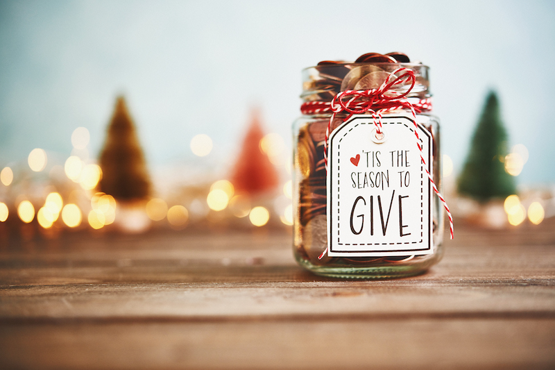 A jar full of pennies with a handwritten label that reads 'Tis the season to give' tied on with festive red and white string