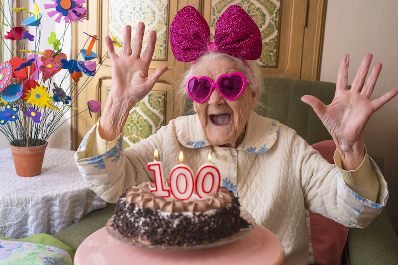 Woman in heart shaped sunglasses and a large bow in her hair laughing behind a 100 year birthday cake