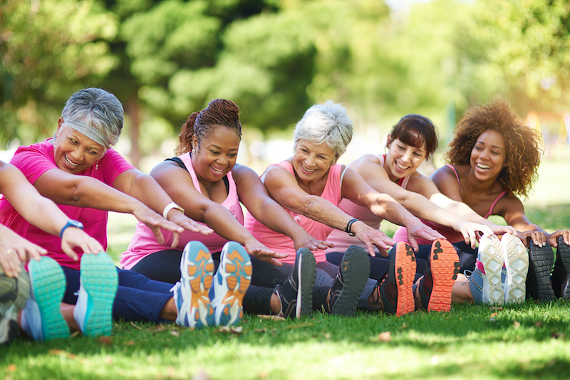 A line of ladies in exercise clothes sitting in the grass at a park stretching their arms out to their toes