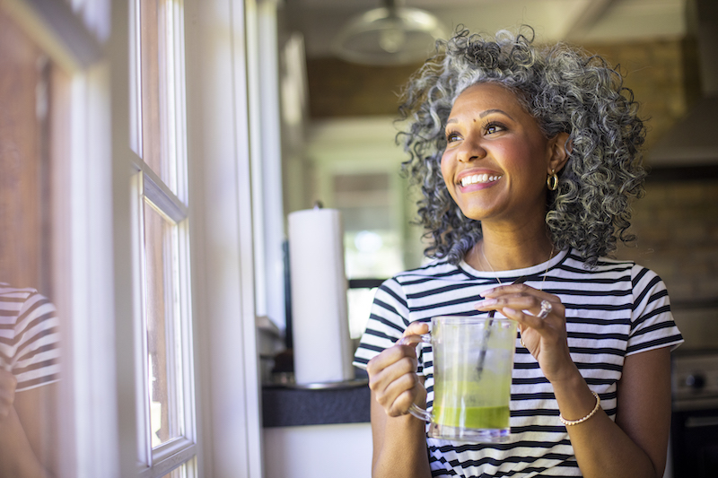 A woman smiling looking out the window while enjoying a healthy green juice drink