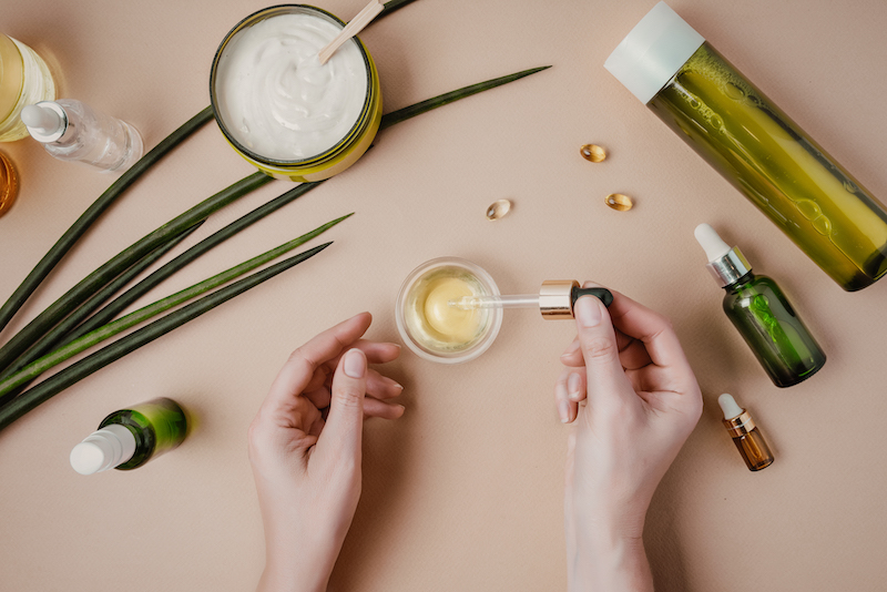 Top-down view of a table with scattered organic green vials and woman's hands using dropper to mix ingredients