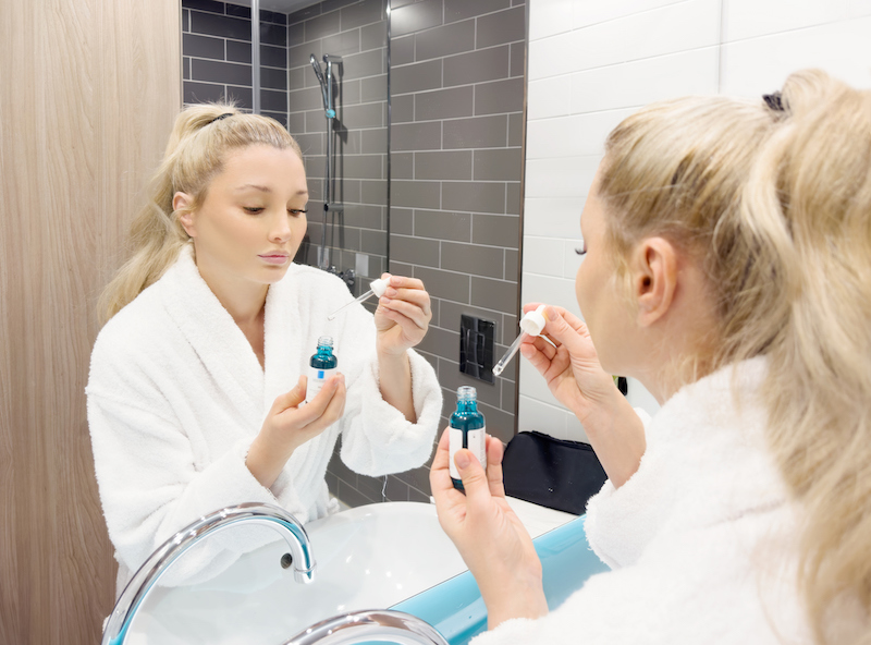 Woman in white robe preparing to apply a skin care product from a blue bottle with a dropper